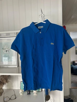 Blå pikétröja från Lacoste - Snygg blå pikétröja från Lacoste med klassisk krage och knappar framtill. Tröjan har korta ärmar och den ikoniska krokodilloggan på bröstet. Perfekt för en avslappnad men stilren look. Storlek S och är sparsamt använd.