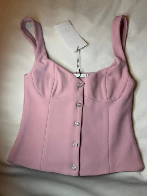 Rosa korsettopp från zara - Supersnygg rosa korsettopp från Zara med breda axelband och dekorativa knappar framtill. Toppen har en figurnära passform och markerade sömmar som ger en feminin siluett. Perfekt att styla med jeans eller kjol för en trendig look.