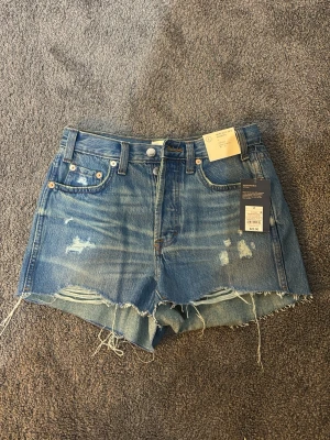 Blå denimshorts med slitningar - Säljer ett par blå denimshorts med råa kanter och slitna detaljer framtill. Klassisk femficksmodell med knapp och dragkedja. Perfekta för en avslappnad och trendig look i sommar.