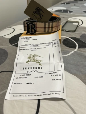 Burberry vändbart bälte med monogram - Säljer ett vändbart Burberry-bälte i skinn med klassiskt beige-randigt mönster på ena sidan och svart på andra. Stort metallspänne med Burberrys monogram. Perfekt accessoar för att lyfta din outfit med lyxig känsla och ikonisk design.