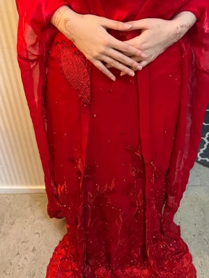 Röd långklänning med paljettdetaljer - Snygg röd långklänning med glittriga paljettdetaljer och broderier. Klänningen har lång ärm och är tillverkad i ett lätt, flödande material som ger en elegant look. Perfekt för dig som vill sticka ut med färg och detaljer.