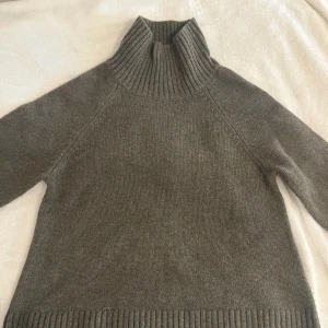 Grå stickad polotröja - Mysig grå stickad polotröja med hög krage och långa ärmar. Tröjan har ribbstickade muddar vid ärmslut, nederkant och krage. Perfekt för kyliga dagar och enkel att matcha med andra plagg.