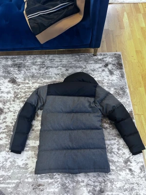 Mörkgrå dunjacka med huva - Snygg mörkgrå dunjacka från The North Face med svarta detaljer på axlar och ärmar. Jackan har två stora framfickor med lock, dragkedja och tryckknappar samt en varm huva. Perfekt för kalla vinterdagar.