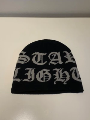 Svart mössa från starlight - Säljer en svart mössa med stor vit text i gotisk stil som täcker hela framsidan. Mössan är mjuk och stretchig, perfekt för kalla dagar och ger en edgy streetwear-vibe. Passar dig som gillar att sticka ut och vill ha en cool accessoar till din outfit.