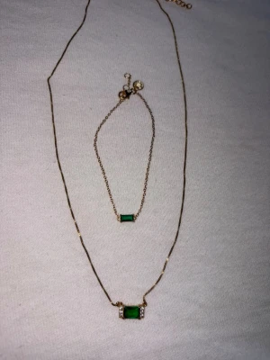 Set med halsband och armband guldgrön ifrån safira - Säljer ett snyggt set med halsband och armband i guldfärgad metall. Båda har en rektangulär grön sten i mitten, omgiven av små klara stenar på sidorna. Kedjorna är tunna och eleganta, perfekta för att lyfta vilken outfit som helst.