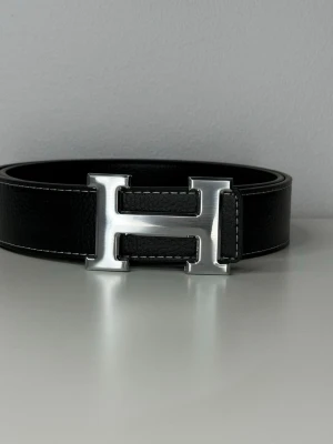 Exklusivt bälte med färgen svart - Stilrent svart bälte med klassiskt H-formad spänne i silverfärgad metall. Skärpet är gjort i exklusivt skinn med diskreta vita sömmar längs kanterna. Perfekt accessoar för att lyfta din outfit.