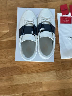 Valentino vita sneakers med Navy blue detalj - Snygga vita sneakers från Valentino med en bred svart läderdetalj över vristen. Klassisk låg modell med rund tå och vita skosnören. Märkeslogga syns på plösen. Skorna är tillverkade i läder och har en stilren, modern design.