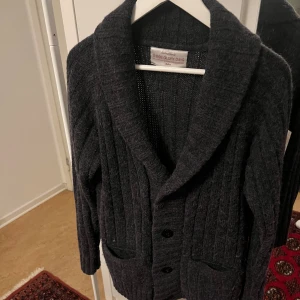 Mörkgrå cardigan - Säljer en mörkgrå stickad kofta från Weekday med sjalkrage och knäppning framtill. Koftan har två fickor och ett grovt ribbstickat mönster som ger en snygg och avslappnad look. Perfekt att slänga på sig över en t-shirt eller skjorta.