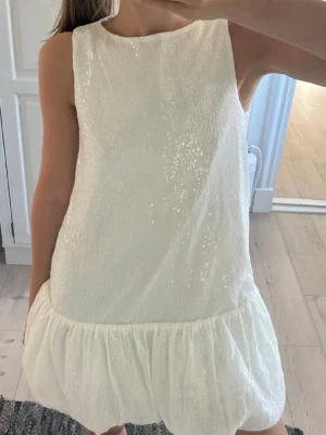 Vit glittrig kort klänning med volang - Supersöt vit kort klänning med glittrigt tyg och volang nertill. Ärmlös modell med rak passform och rund halsringning. Perfekt för dig som vill sticka ut lite extra! Materialet känns lätt och skimrande. 