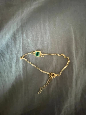 Guldigt armband med grön sten - Snyggt armband i guldfärgad metall med en fyrkantig grön sten i mitten, omgiven av små detaljer. Kedjan är tunn och har justerbar längd med karbinlås. Perfekt för dig som gillar stilrena accessoarer med en färgklick.