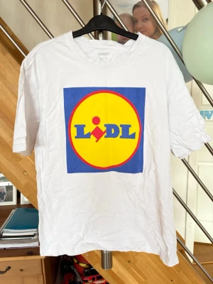 Vit Lidl t-shirt från Livergy - Vit t-shirt från Livergy med stort Lidl-tryck på bröstet i blått, gult och rött. Klassisk rund hals och korta ärmar. Perfekt för dig som gillar att sticka ut med humor och enkel stil. Gjord i mjuk bomull som känns skön mot huden.