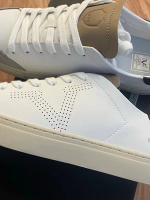 19.69 italia sneakers  - Stilrena vita sneakers från Valsport Milano med perforerat V på sidan, beige och mörkblå detaljer samt grå mocka vid tån. Skorna har vita skosnören och en klassisk låg profil. Tillverkade i läder och mocka för en clean look.