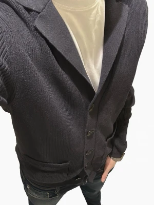 Cardigan Dressmann - Snygg marinblå stickad Cardigan från Dressmann med knäppning framtill och två fickor. Koftan har krage och ribbade muddar vid ärmslut och nederkant. Perfekt att slänga över en t-shirt för en chill och stilren look🤩 Inte använd mycket, blir pösig i armarna när man inte tvättar den på ett tag men drar till sig när man tvättar. Nypris 800kr. Pris kan diskuteras vid smidig affär. Skickar snabbt📦