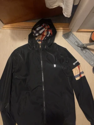 Svart vindjacka från Burberry - Snygg svart vindjacka från Burberry med klassiskt rutigt foder i huvan och på ena ärmen. Jackan har dragkedja framtill, huva och diskret Burberry-logga på bröstet. Storlek M passar även L 