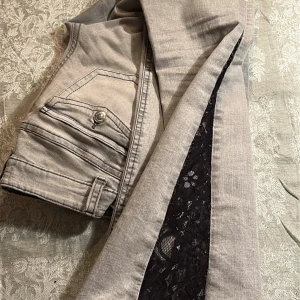 Grå jeans med spetsdetalj - Snygga grå jeans med unik svart spetsdetalj längs sidan av benen. Jeansen har klassiska fickor med knapp och dekorativa sömmar. Materialet är mjukt och stretchigt, vilket ger en bekväm passform. Perfekta för dig som vill sticka ut med en cool twist på vanliga jeans. Omsydda med spets.