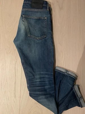 Mörkblå Levis jeans selvedge - Snygga mörkblå slim fit jeans med slitningar och vikta benslut. Jeansen har klassiska fem fickor och en skinnpatch i midjan. Perfekta för en trendig och avslappnad look. W30 L32