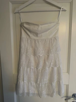 Vit spetsklänning utan axelband  - Söt vit kortklänning med vackra spetsdetaljer i lager. Klänningen har en dragkedja i ryggen och är axelbandslös upptill. Perfekt för sommarens alla tillfällen.