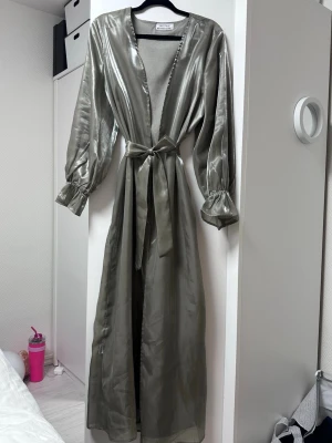 Lång olivgrön satin-klänning med bälte - Supersnygg långklänning i glansig olivgrön satin. Klänningen har långa ärmar med resår vid ärmslut, v-ringad urringning och ett matchande bälte i midjan. Perfekt för dig som vill sticka ut med en elegant look.