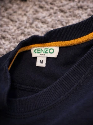 Svart sweatshirt från Kenzo - En stilren svart sweatshirt från Kenzo i storlek M. Tröjan har rund halsringning och ribbade muddar vid ärmslut och nederkant. Tillverkad i mjukt material som känns skönt mot huden. Perfekt för en clean och avslappnad look.