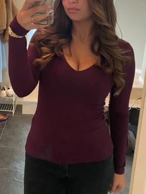Vinröd långärmad v-ringad topp - Vinröd långärmad topp med v-ringning och figurnära passform. Perfekt att styla med jeans eller kjol för en enkel men snygg look. Mjuk och stretchig känsla, passar dig som gillar stilrena basplagg.