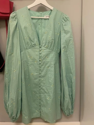 Mintgrön klänning från Adoore - Supersöt mintgrön klänning från Adoore med små gula blommor, v-ringning och klädda knappar framtill. Klänningen har långa puffärmar och lätt markerad midja. Materialet ser ut att vara bomull eller bomullsblandning. Perfekt för dig som gillar romantisk och trendig stil.