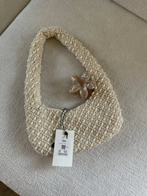 Pärlprydd handväska från Bik Bok - Superfin handväska från Bik Bok i beige med pärlbroderier över hela väskan. Väskan har en unik, mjukt trekantig form och dragkedja upptill. Perfekt accessoar för att lyfta vilken outfit som helst. Materialet är tyg täckt med pärlor för en lyxig känsla.
