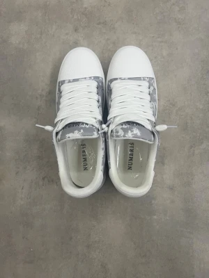 Vita och grå sneakers från NUMERIS - Snygga sneakers från NUMER'S i vitt och grått med coolt kamouflagemönster på sidorna. Skorna har vita, breda skosnören och en klassisk låg siluett. Ovandelen är i textil och syntet, och sulan är vit och räfflad för extra grepp.