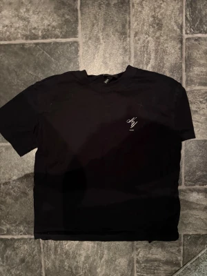 Svart t-shirt med vit text från NY - Säljer en svart t-shirt från NY med vit text och logga på bröstet samt stor vit text på ryggen där det står 'NY' och 'S/S 20XX'. Klassisk passform och rund halsringning. Perfekt för dig som gillar stilrena och enkla plagg.