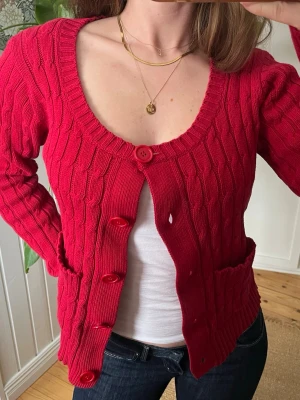 Röd kabelstickad kofta med fickor - Snygg röd kabelstickad kofta med stora knappar samt fickor. 100% bomull. Står L men den är gammal ich passar perfekt på mig som bär storlek 36. 