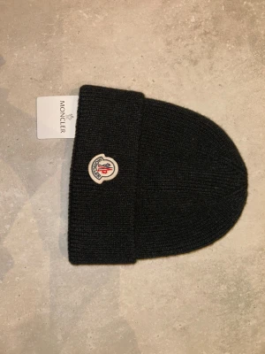 Svart ribbstickad mössa från Moncler - Snygg svart ribbstickad mössa från MC med uppvikt kant och klassisk MC logga framtill. Tillverkad i mjukt material som håller dig varm och stilren under kalla dagar. Perfekt accessoar för dig som vill ha både komfort och trendig look.