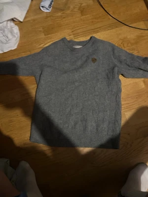 Grå kabelstickad tröja Lexington - Snygg grå kabelstickad tröja från Lexington med rund halsringning och diskret logomärke på bröstet. Tröjan har ribbstickade muddar vid ärmslut och nederkant, perfekt för en stilren och klassisk look.