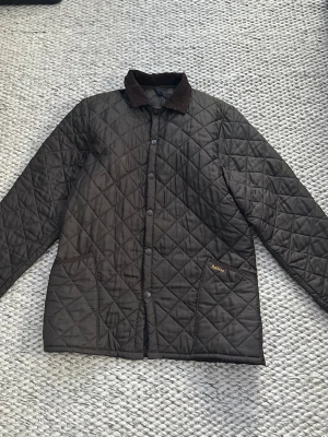 Brun quiltad jacka från Barbour - Brun quiltad jacka från Barbour med krage i manchester och tryckknappar framtill. Jackan har två stora fickor och diskret Barbour-logga på ena fickan. Storlek M, men stor i storleken. Mått: Bröstvidd: 58cm, Längd: 80cm, Armlängd 65cm.