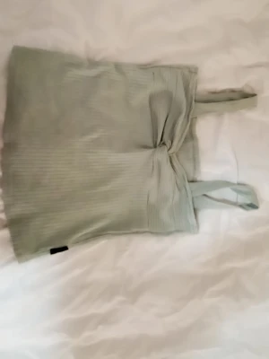 Ljusgrön ribbad linnetopp med knyt - Trendig ljusgrön linnetopp med ribbad struktur och snygg knytdetalj framtill. Toppen har smala axelband och croppad passform, perfekt till jeans eller kjol. Enkel men ändå stilren design som passar till många olika looks.