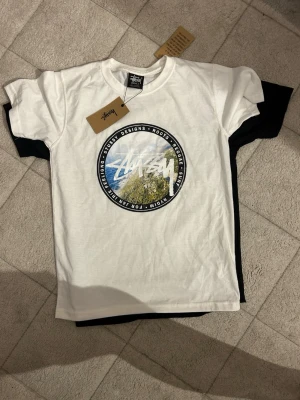 Stüssy t-shirt pack - Två limited edition t-shirts från Stüssy . Ena är vit med rund print framtill och Stüssy-logga, den andra är svart med stort grafiskt tryck och Metallica-motiv på ryggen. Båda har klassisk passform och är i mjuk bomull. Perfekt för dig som gillar streetwear och bandmerch.8 tishor ingår 