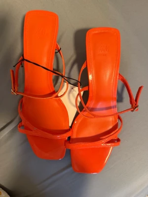 Oranga sandaletter med remmar H&M - Säljer ett par trendiga oranga sandaletter från H&M med smala remmar och fyrkantig tå. Sandalerna har en liten klack och justerbart spänne vid ankeln. Tillverkade i syntetmaterial med glansig finish. Perfekta för att lyfta vilken outfit som helst.
