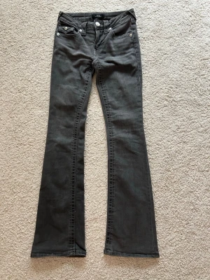 Mörk gråa bootcut jeans från true religon  - Säljer ett par svarta bootcut jeans från True religon med klassisk femficksdesign och silverfärgade knappar. Jeansen har fickor med lock bak och är tillverkade i mjukt denim-material med stretch. Det är använda en gång, säljer pga för små.