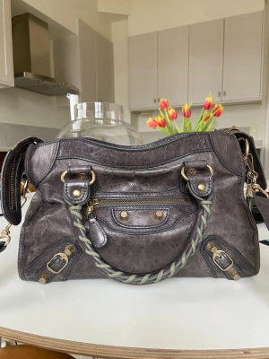 Balenciaga City väska i mörkgrått läder - Balenciaga City handväska i mörkgrått/brunt läder med guld detaljer. Väskan har både handtag och axelrem. Köpt på Vinted där säljaren sa att hon hade köpt den på en butik för designväskor. Mått: 30,20, 10 cm. Pris kan diskuteras vid snabb affär. 