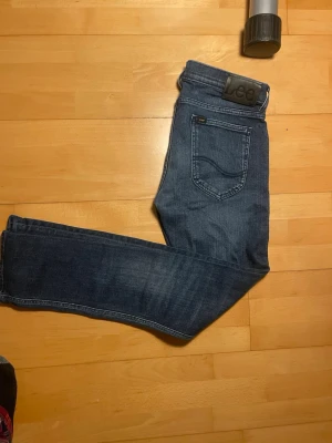 Mörkblå Lee jeans med smal passform - Snygga mörkblå jeans från Lee med klassisk femficksdesign och smal passform. Jeansen har subtila slitningar och Lee-logga på bakfickan samt läderpatch i midjan. Tillverkade i mjukt denimtyg som sitter skönt hela dagen. Skriv vid minsta fundering riktigt snygga fades