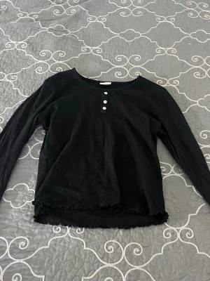 Svart långärmad topp med knappar - En svart långärmad topp med rund hals och tre vita knappar framtill. Lite genomskinlig o kort. Toppen har en lätt vågig kant nertill och är gjord i mjukt bomullstyg. Enkel och stilren design som passar till mycket.