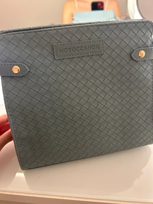 Grå flätad handväska från Moroccanoil - Snygg grå handväska/ necessär från Moroccanoil med flätad struktur och guldfärgade detaljer. Väskan har en fyrkantig form och är tillverkad i konstläder. Perfekt för dig som gillar stilrena accessoarer med en twist.  2 för 300 1 för 200💕