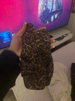 Leopardmönstrade slip-in sandaler - Säljer ett par trendiga slip-in sandaler med leopardmönster. Sandalerna har fyrkantig tå och låg klack, samt breda band över foten. Materialet ser ut att vara textil med mjuk finish. Perfekta för dig som vill sticka ut med ett djurmönster.