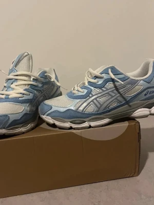 Blåvita sneakers från Asics - Snygga sneakers från Asics i ljusblått och vitt med meshpaneler och detaljer i syntet. Skorna har snörning och en bekväm, platt sula. Perfekta för dig som gillar sportig stil och vill ha något som sticker ut pris kan diskuteras .