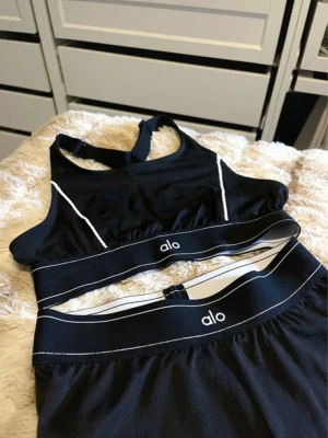 Svart träningsset från Alo - Snyggt svart träningsset från Alo med sport-bh och tights. Setet har vita kontrastsömmar och bred resår med Alo-logga. Materialet är stretchigt och passar perfekt för gym, yoga eller löpning. Sport-bhn har brottarrygg och tightsen har hög midja.
