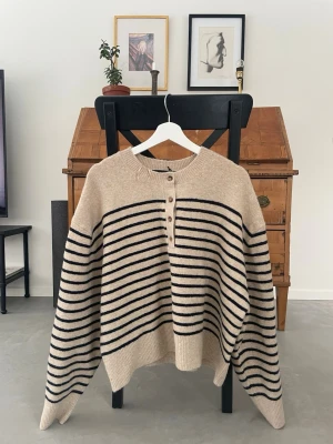Beige stickad tröja med knappar från Zara - Säljer en beige stickad tröja med svarta horisontella ränder och fyra knappar framtill. Tröjan har rund halsringning och ribbade muddar vid ärmslut och nederkant. Oversized passform och mjukt material, perfekt för kyliga dagar.