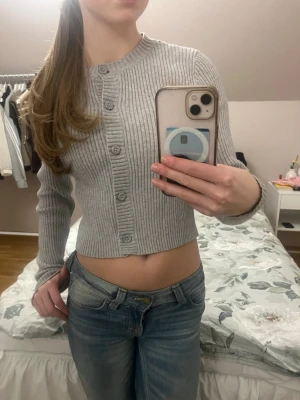 Grå ribbad cropped kofta - Säljer en ljusgrå ribbstickad cropped kofta med rund hals och knäppning framtill. Koftan har långa ärmar och sitter tajt mot kroppen, perfekt att styla över ett linne eller t-shirt. Enkel och stilren design som passar till det mesta.