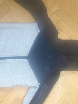 Nike Tech Fleece hoodie svart/grå - Nike Tech Fleece hoodie i svart och ljusgrått med dragkedja framtill och huva. Snygg tvåfärgad design och diskret Nike-logga på bröstet. Tillverkad i mjukt och bekvämt material, perfekt för chill eller träning. Fickor med dragkedja på sidorna för smart förvaring.