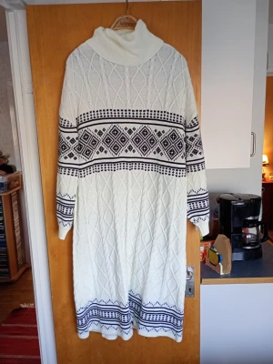 Vit stickad långklänning med mönster - Vit stickad långklänning med svart geometriskt mönster och polokrage. Klänningen har lång ärm och är perfekt för kyligare dagar. Snygg strukturstickning och mjukt material som känns skönt mot huden.