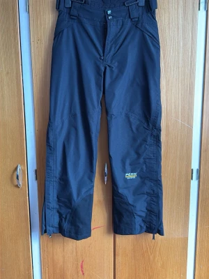 Svarta täckbyxor Peak Performance - Snygga svarta täckbyxor från Peak Performance med GORE-TEX XCR-material som står emot både vind och vatten. Byxorna har dragkedjor vid bensluten och förstärkta partier för extra hållbarhet. Perfekta för vinterns alla äventyr.