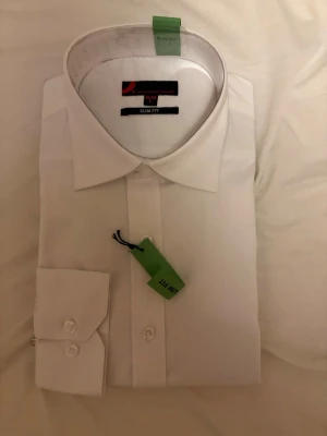 Vit slim fit skjorta från Dressmann - Klassisk vit skjorta från Dressmann i slim fit-modell. Skjortan har lång ärm, knappar framtill och en stilren, tidlös design som passar till många olika looks. Perfekt för dig som gillar en smalare passform och enkel, clean stil.