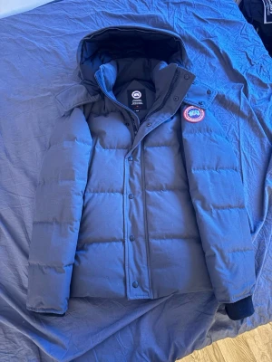 Mörkblå dunjacka från Canada Goose - Säljer en mörkblå dunjacka från Canada Goose med avtagbar huva och klassisk logopatch på ärmen. Jackan har en puffig siluett, dragkedja och knappar framtill samt två stora fickor. Perfekt för kalla vinterdagar och riktigt varm. Haft jackan lite över ett år. Digitalt kvitto finns på jackan och ja allt som visar att den är äkta. Jackan är så god som ny finns knappt en skråma på den. Skriv om du undrar något !